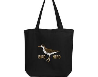 May include: Black tote bag featuring a bird illustration with the text "BIRD NERD" in a brown font. The bird is brown, white, and black. The bag has two black handles and is hanging from a black hook.