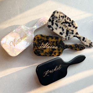 Op de afbeelding: Vier gepersonaliseerde haarborstels in verschillende kleuren en patronen. Eén is parelmoerwit met "Victoria" in gouden script, een andere is zwart-wit met "Marissa", een schildpad met "Alaina" en een zwarte met "Rachel".