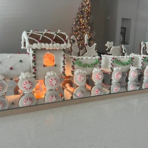 May include: A gingerbread train with a lit window in the engine car. The train is decorated with white icing, red and green sweets, and green garland. The train is sitting on a mirrored surface.