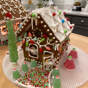 May include: A decorated gingerbread house with white icing, colorful candy lights, and a Hershey's Kiss on top. The house sits on a clear, ridged plate, surrounded by gumdrop trees and a path of sprinkles. A second gingerbread house is visible in the background.