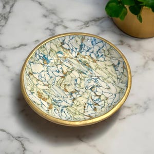 May include: A small, white ceramic bowl with a gold rim and a blue and green marbled design. The bowl is on a white marble surface.