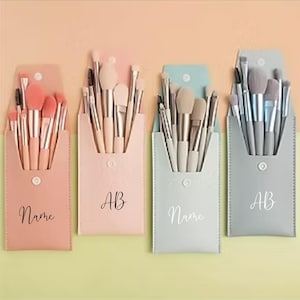 Op de afbeelding: Vier make-up kwastensets in pastelkleurige etuis. Elke etui bevat meerdere kwasten en heeft een knoopsluiting. Twee etuis hebben de initialen "AB", en twee het woord "Name".