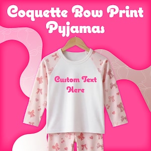 May include: A long-sleeve pajama set featuring a white center panel with the text Custom Text Here in pink, surrounded by light pink sleeves and pants patterned with small pink bows and hearts. The top is displayed on a wooden hanger.