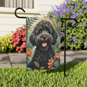 Puede incluir: Un caniche negro con la lengua rosa que sobresale está rodeado de flores naranjas y verdes. El perro está mirando directamente a la cámara. La imagen es una bandera de jardín.