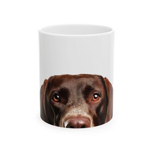 May include: A white ceramic mug with a brown and white dog's face peeking over the rim. The dog has brown eyes and a brown nose.
