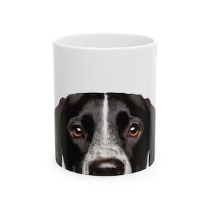 May include: White ceramic mug with a black and white dog's face peeking over the rim. The dog has brown eyes and a black nose.