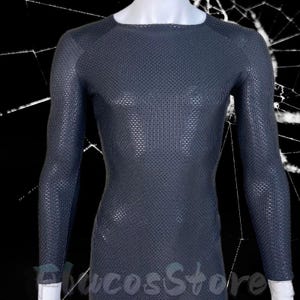 Może przedstawiać: Ciemnoszara koszulka z długim rękawem i teksturowanym wzorem w kratkę. Koszulka ma okrągły dekolt i jest prezentowana na manekinie. Widoczny jest dół koszulki, a rękawy sięgają do nadgarstków. Koszulka jest wykonana z elastycznego materiału.