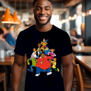 May include: Black t-shirt with a cartoon group design. The cartoon characters are depicted in a variety of poses and clothing, with a central character wearing a red shirt and blue trousers. The design is bright and colourful.