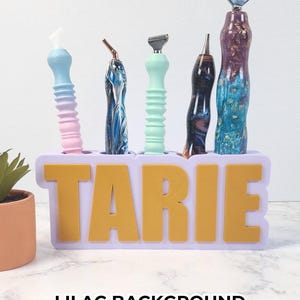 May include: A lilac-colored holder with gold lettering that spells "TARIE" holds five razors with colorful handles. The razors have blue, pink, green, and marbled designs. The text "LILAC BACKGROUND WITH GOLD LETTERING" is at the bottom.