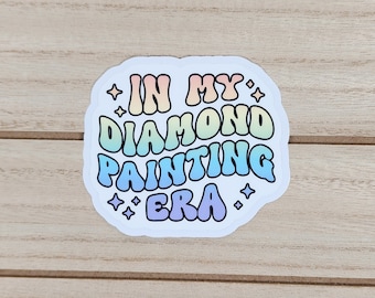 以下が含まれることがあります： テキストの周りに星が散りばめられた、「In My Diamond Painting Era」と書かれた虹色のグラデーションデザインの白いステッカーです。