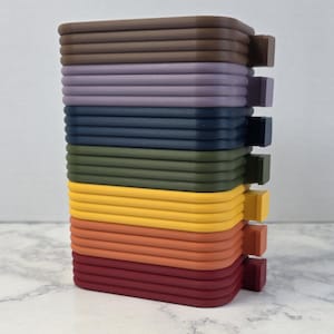 May include: A stack of seven rectangular, colorful, and textured objects. The colors include brown, purple, navy, olive green, yellow, orange, and red. Each object has a small, square tab on one side.