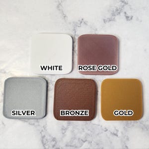 May include: Five square color swatches in white, rose gold, silver, bronze, and gold. Each swatch has rounded corners and a textured surface. The color names are printed in black, centered on each swatch.