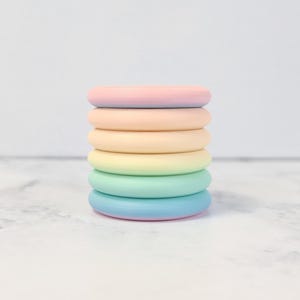 Op de afbeelding: Een stapel van zes pastelkleurige siliconen ringen, elke ring is een andere tint roze, geel, groen en blauw. De ringen zijn op elkaar gestapeld, waardoor een regenboog effect ontstaat.
