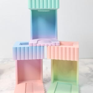 Puede incluir: Cuatro soportes para teléfono de silicona de forma cuadrada, con efecto degradado, de color pastel. Los soportes tienen un borde festoneado y un diseño texturizado y acanalado. Los soportes están apilados uno encima del otro, con el soporte superior de color azul claro y verde, el segundo soporte de color morado claro y rosa, el tercer soporte de color rosa claro y naranja, y el soporte inferior de color verde claro y amarillo.