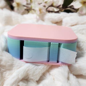 May include: A pastel pink, blue, and green tape dispenser with a white tape roll. The dispenser has a layered design with a rounded top.
