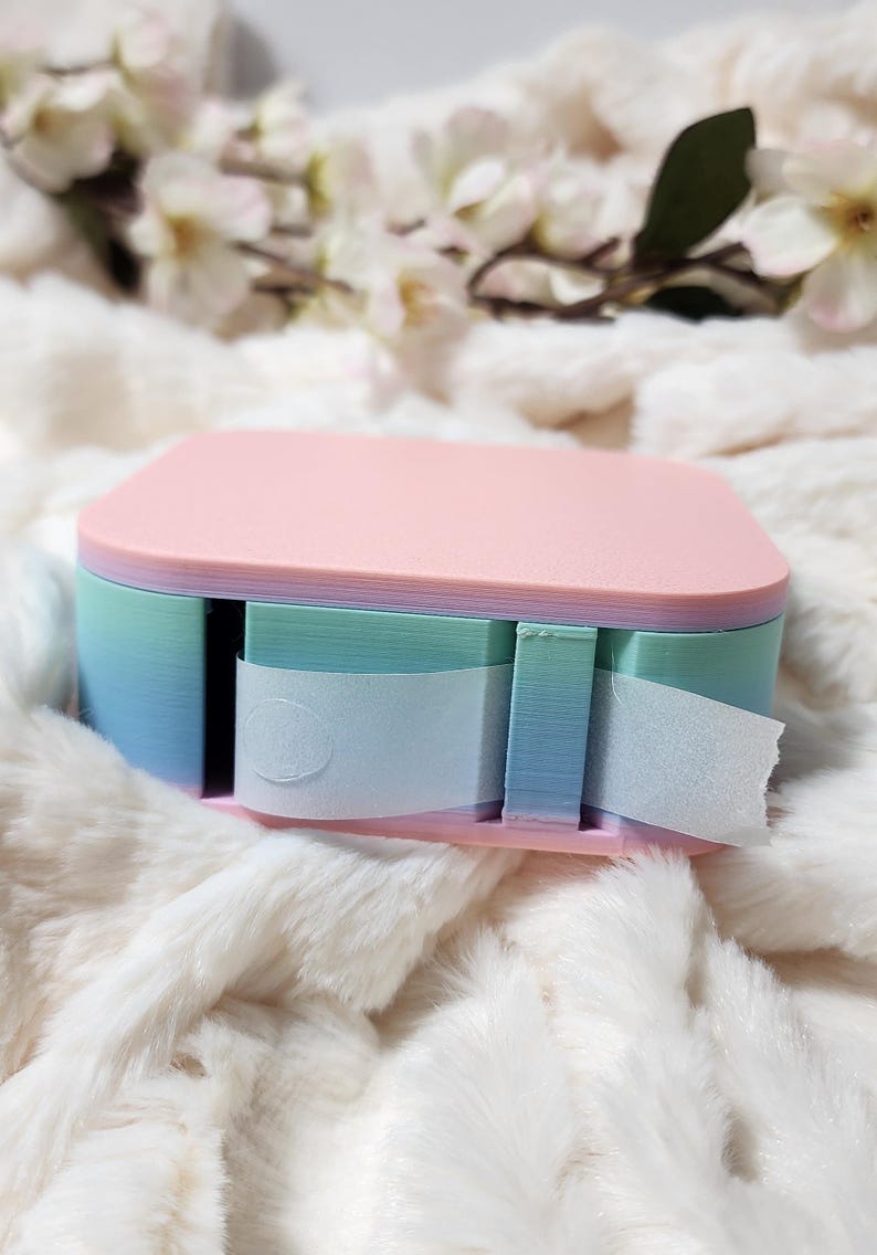 May include: A pastel pink, blue, and green tape dispenser with a white tape roll. The dispenser has a layered design with a rounded top.