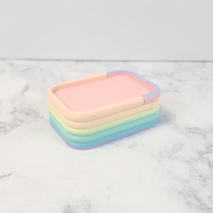 May include: A stack of five rainbow-colored silicone phone stands. The stands are in shades of pink, orange, yellow, green, and blue.