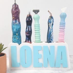 May include: A collection of five decorative tools with colorful handles, displayed in a white holder with the name "LOENA" in sky blue letters. The tools have various designs and colors, including blue, purple, and green.