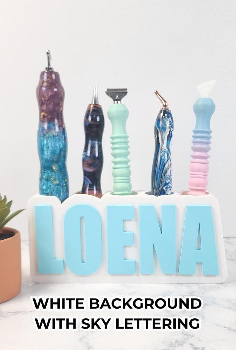 May include: A collection of five decorative tools with colorful handles, displayed in a white holder with the name "LOENA" in sky blue letters. The tools have various designs and colors, including blue, purple, and green.