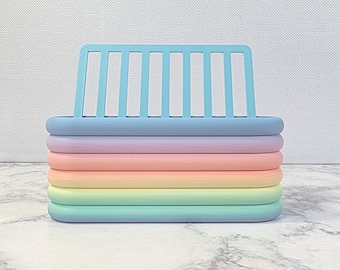 May include: A pastel-colored desk organizer with a light blue slotted back and a stack of rounded layers in shades of blue, purple, pink, yellow, and green. The organizer sits on a marble-patterned surface.