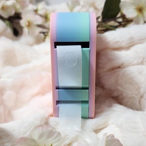 Peut inclure: Un distributeur de ruban adhésif rose pastel, bleu et vert avec un rouleau de ruban blanc. Le distributeur est posé sur une surface blanche et moelleuse.