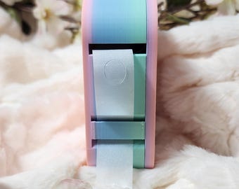 May include: A pastel pink, blue, and green tape dispenser with a roll of white tape. The dispenser is sitting on a white fluffy surface.