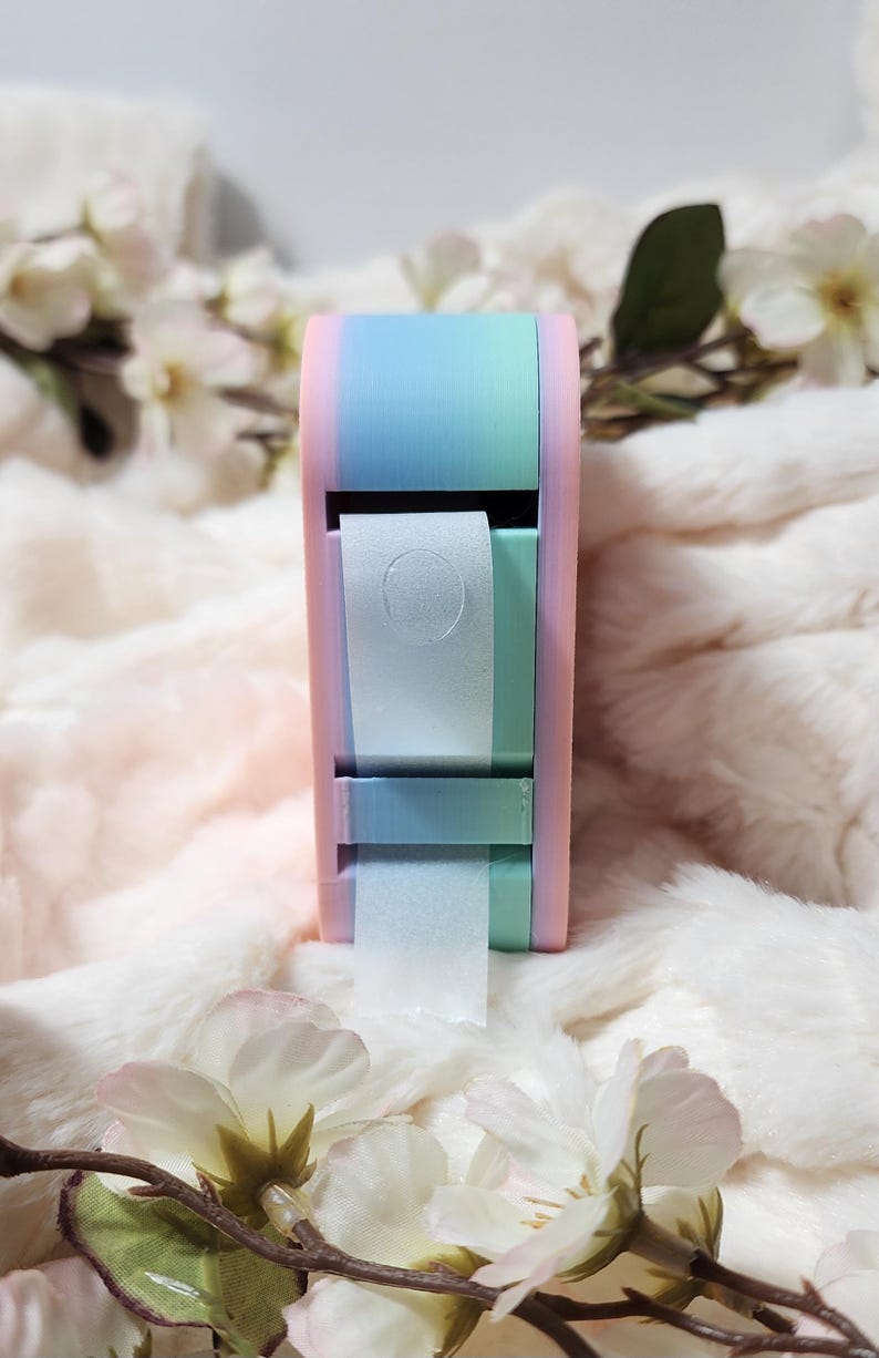 May include: A pastel pink, blue, and green tape dispenser with a roll of white tape. The dispenser is sitting on a white fluffy surface.