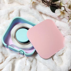 May include: A pastel blue, pink, and white square container with a roll of clear tape inside. The container has a lid that is a solid pink color.