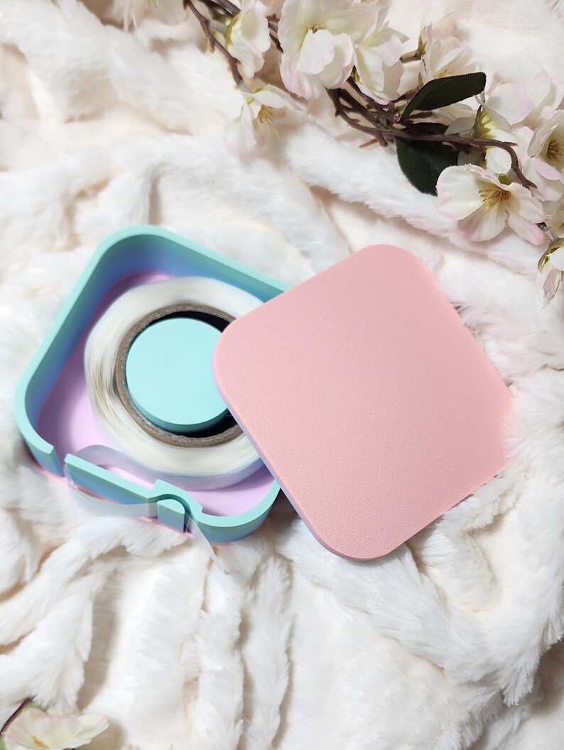 May include: A pastel blue, pink, and white square container with a roll of clear tape inside. The container has a lid that is a solid pink color.