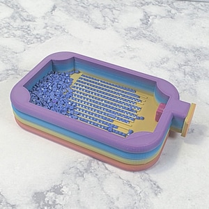 Puede incluir: Una bandeja multicolor de varias capas con forma rectangular, diseñada para pintura con diamantes. La bandeja contiene numerosas cuentas pequeñas azules en forma de diamante. La bandeja tiene un interior amarillo claro y un exterior morado con un pequeño botón rosa.