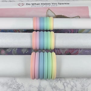 Puede incluir: Tres lienzos enrollados de pintura de diamantes están asegurados con bandas de silicona de colores pastel arcoíris. Los lienzos son blancos y presentan diseños de arte de diamantes coloridos. El texto "Do What Makes You Sparkle" es visible.