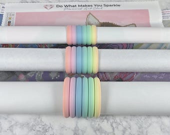 May include: Three rolled diamond painting canvases are secured with pastel rainbow-colored silicone bands. The canvases are white and feature colorful diamond art designs. The top canvas has the text "Do What Makes You Sparkle".