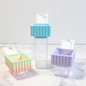 May include: Three small clear plastic containers with colorful lids. The lids are removable and have a ribbed texture. The containers are empty and are arranged in a row on a white marble surface.