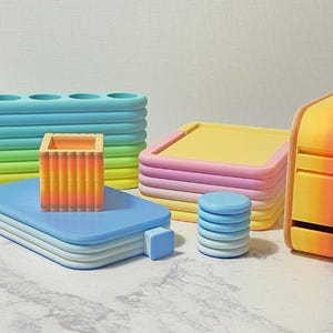 Op de afbeelding: Een verzameling kleurrijke, geometrische bureau-organizers. Inclusief een blauw-groene pennenhouder, een blauw dienblad, een stapel roze en gele trays, een kleine blauwe stapel en een geel-oranje dispenser.