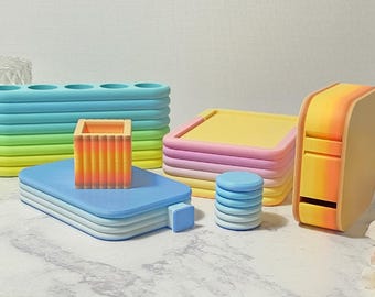 May include: A collection of colorful, geometric desk organizers. Includes a blue and green pencil holder, a blue tray, a stack of pink and yellow trays, a small blue stack, and a yellow and orange dispenser.