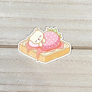 Op de afbeelding: Een sticker met een slapende beer op een toast met aardbeienjam en een grote aardbei. De beer is crèmekleurig, de aardbei is roze met witte stippen en de toast is goudbruin.