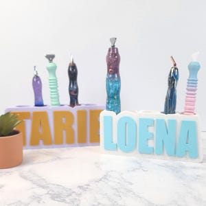 May include: Two personalized pen holders with pens. One holder is lavender with yellow letters spelling "TARIE", the other is white with blue letters spelling "LOENA". Pens are various colors.