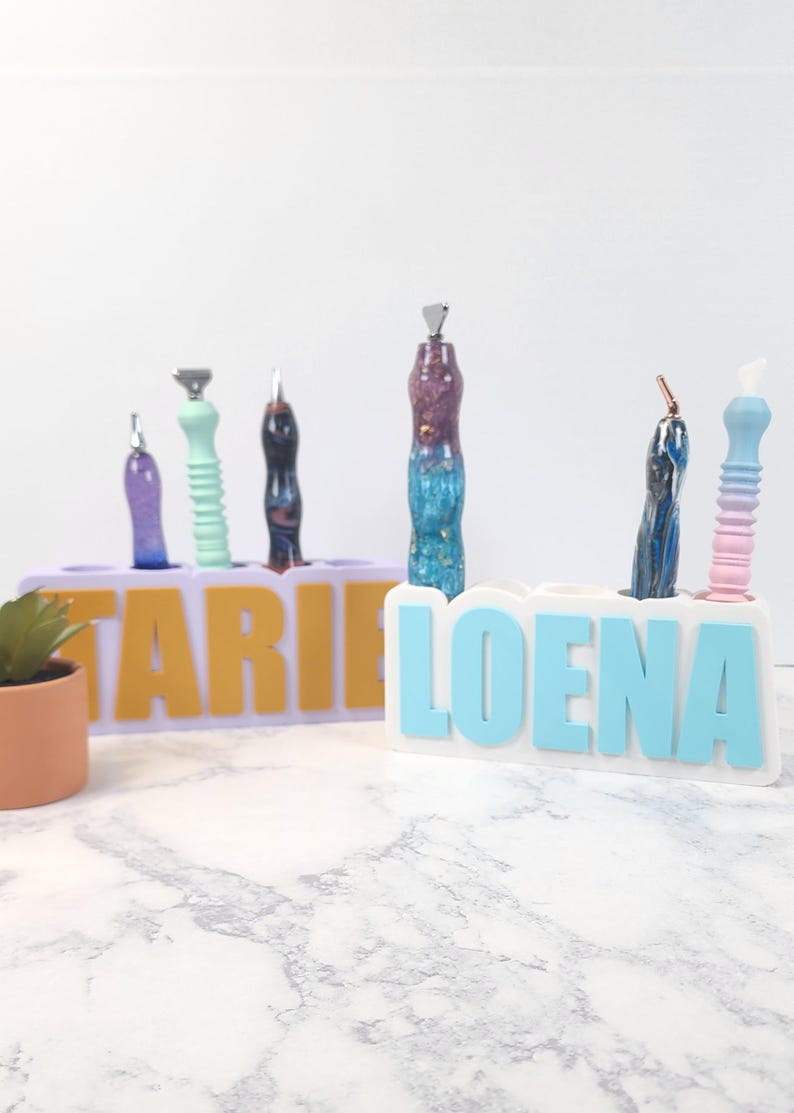 May include: Two personalized pen holders with pens. One holder is lavender with yellow letters spelling "TARIE", the other is white with blue letters spelling "LOENA". Pens are various colors.