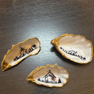 Può includere: Tre conchiglie di ostriche dipinte in oro con silhouette nere della scena della natività. Le conchiglie sono disposte su una superficie di legno scuro.