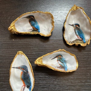 Op de afbeelding: Vier oesterschelpen met goud geschilderde randen, elk met een geschilderde afbeelding van een ijsvogel. De vogels hebben blauwe, oranje en zwarte veren. De schelpen liggen op een donkerbruin oppervlak.