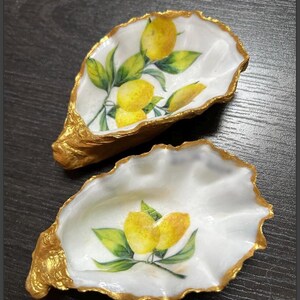 以下が含まれることがあります： 金色の縁取りが施された白いオイスターシェル2つ。レモンの枝のデザインが描かれています。シェルは、ジュエリーや小物などの小さなアイテムを入れるのに最適です。