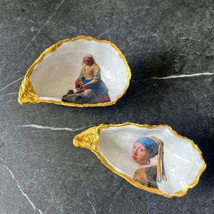 Puede incluir: Dos conchas de ostra decorativas. Las conchas tienen un borde dorado y un interior blanco. Una concha presenta una impresión de una mujer vertiendo de una jarra, y la otra una mujer con un pañuelo azul.