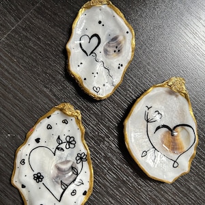 Op de afbeelding: Drie oesterschelpen met gouden rand, elk beschilderd met een ander ontwerp met een hart en andere details. De schelpen zijn wit met zwarte inkttekeningen.
