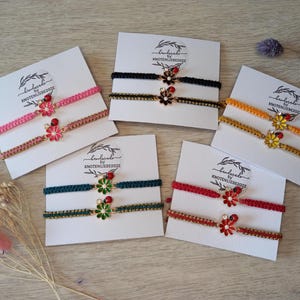 Könnte beinhalten: Eine Auswahl handgefertigter Armbänder in verschiedenen Farben wie Rosa, Schwarz, Orange, Grün und Rot. Jedes Armband hat einen Blumenanhänger mit roten und gelben Akzenten. Die Armbänder werden auf weißen Karten mit dem Text "handmade by KNOTENLIEBE2022" präsentiert.