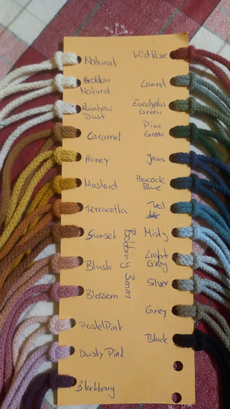 May include: A color chart featuring various shades of cord, with names like "Natural," "Blossom," and "Blackberry." The cords are attached to a yellow card with handwritten color names. The cords are 3mm thick.