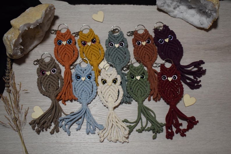 May include: A collection of macrame owl keychains in various colors, including brown, orange, yellow, blue, and green. Each owl has a silver keyring and a tassel tail. The owls are decorated with small beads for eyes and a heart-shaped beak.