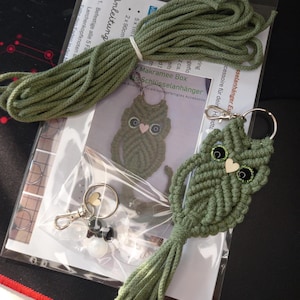 Könnte beinhalten: Ein Makramee-Eulen-Schlüsselanhänger-Set in Grün. Das Set enthält grüne Kordel, einen Schlüsselanhängerring, Perlen und eine Anleitung. Die fertige Eule hat schwarze Augen, ein Holzherz und eine Quaste.