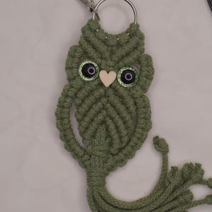 May include: A handmade macrame owl keychain in olive green. The owl has large, glittery eyes and a small wooden heart for a beak. A silver-tone clasp and key ring are attached to the top. The owl's body has a tassel at the bottom.
