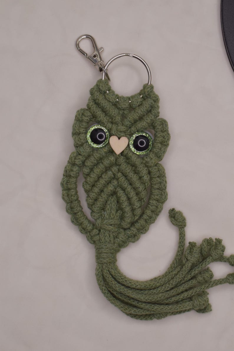 May include: A handmade macrame owl keychain in olive green. The owl has large, glittery eyes and a small wooden heart for a beak. A silver-tone clasp and key ring are attached to the top. The owl's body has a tassel at the bottom.