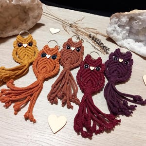 May include: A collection of handmade macrame owl keychains in various warm colors, including mustard yellow, orange, and burgundy. Each owl has a silver keyring, black eyes, and a small wooden heart. The keychains are displayed on a light wooden surface.